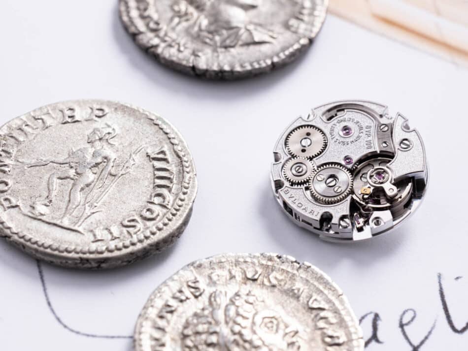 Vista macro del calibre Piccolissimo BVP100, desarrollado por Bvlgari en Le Sentier, comparado con auténticas monedas romanas del periodo 198–297 d.C
