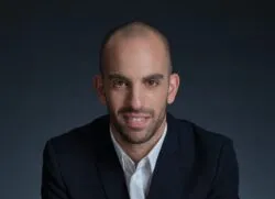 Portrait of a man in a navy blazer and white shirt, seated with arms crossed on a dark table, wearing a silver wristwatch.