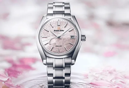 Close-up of a silver Grand Seiko wristwatch with a pink textured dial and date window, set against a soft pink floral background.