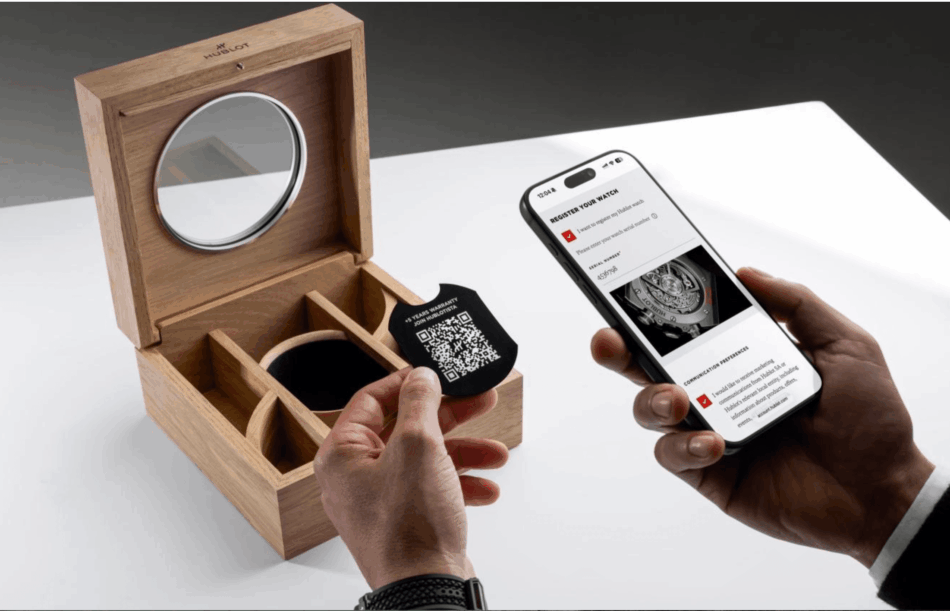 HUBLOT GARANTÍA 5+5 Watches World Open wooden watch box with a round mirror a hand holds a QR code tag beside a smartphone showing watch registration