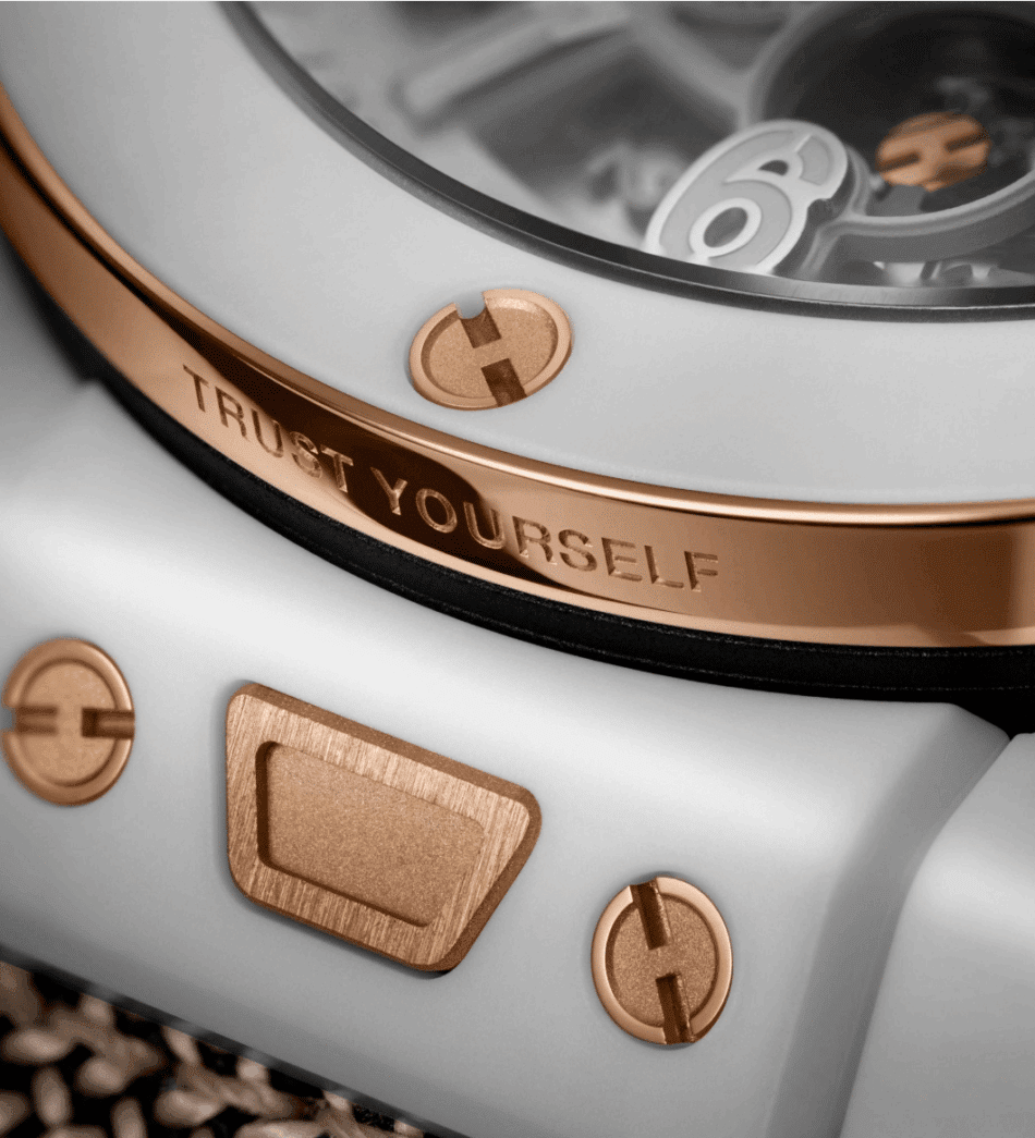 HUBLOTReloaded Kylian Mbappé White CeramicDETALLE Watches World Close up of a light gray device with rose gold screws and a engraved 'TRUST YOURSELF' text on a metal rim, showing a glossy finish.