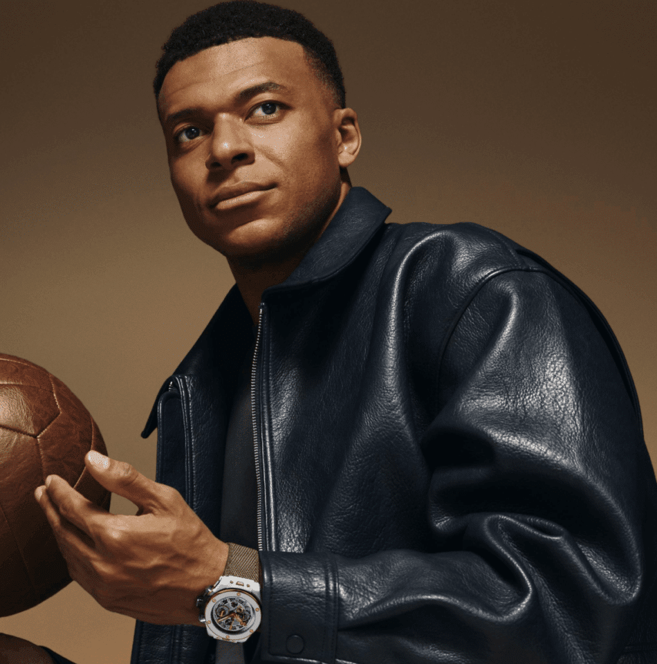 HUBLOTReloaded Kylian Mbappé White CeramicKYLIAN Watches World Portrait of a man in a dark blue leather jacket holding a brown vintage football against a tan backdrop