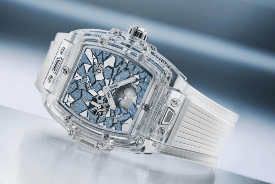 HUBLOTThe Spirit Of Big Bang ImpactCOMPLETO Watches World Transparent square wristwatch with a clear case blue mosaic dial and white strap resting on a reflective surface showcases mechanical inner workings