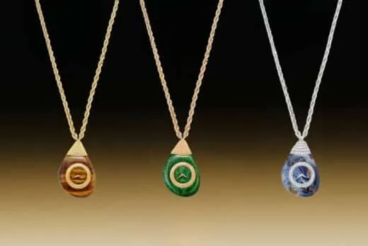 Tres colgantes de piedras preciosas en forma de lágrima con cadenas de oro que cuelgan sobre un fondo con un degradado de negro a marrón (naranja, verde, azul).
