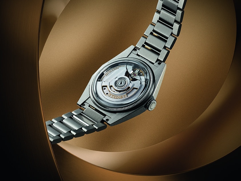 Así es el TUDOR Monarch diseño facetado y calibre de manufactura Watches World Reloj de pulsera de acero inoxidable con mecanismo mecánico a la vista fotografiado sobre un fondo de color bronce cálido