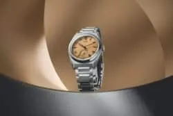 Reloj de pulsera Tudor de acero inoxidable con carátula beige, expuesto sobre un fondo metálico curvado.