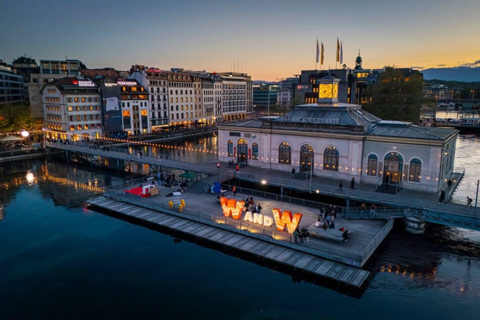 WWG26251InthecityDay3 Watches World Watches and Wonders Geneva 2026 Orange flag with white text flutters in wind at an outdoor rally with white flags and a Swiss flag in the cloudy city background behind itSunset over a bustling riverside city with illuminated storefronts along a canal and a large illuminated W sculpture on the quay