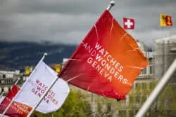Watches and Wonders Geneva 2026 Orange flag with white text flutters in wind at an outdoor rally, with white flags and a Swiss flag in the cloudy city background behind it.