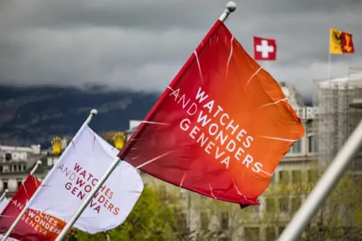 Watches and Wonders Geneva 2026 Orange flag with white text flutters in wind at an outdoor rally, with white flags and a Swiss flag in the cloudy city background behind it.