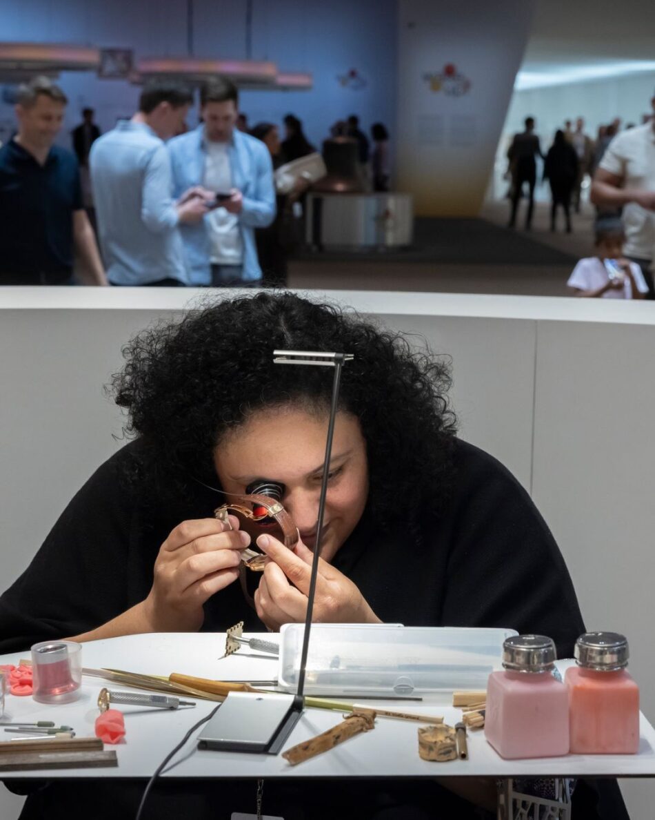Watches and Wonders Geneva 2026 3 Watches World Watches and Wonders Geneva 2026 Curly haired woman examines a wristwatch with a loupe at a white workbench tools and pink bottles nearby