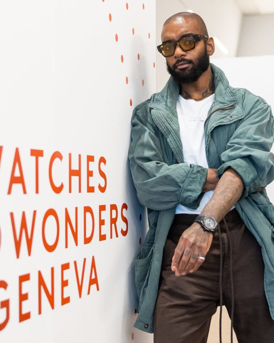 Watches and Wonders Geneva 2026 9 Watches World Watches and Wonders Geneva 2026Man with a beard and sunglasses leans against a white wall with orange text reading 'WATCHES WONDERS GENEVA'.