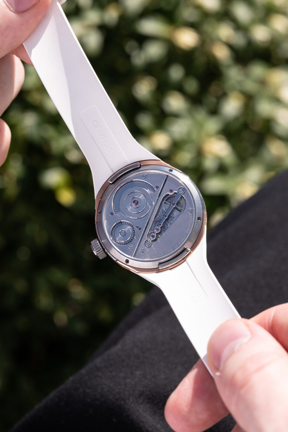 2603dominiquerenaudwatch60 Watches World Close up of a white watch strap being held exposing the back of a mechanical wristwatch with visible gears and rotor