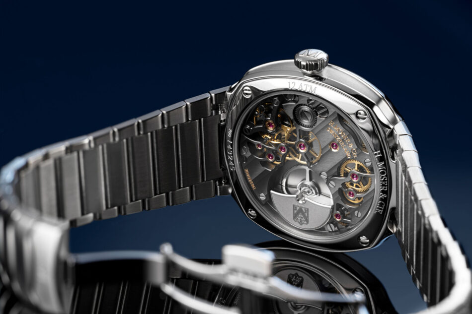 H Moser Streamliner Small Seconds Lime Green ATRAS COMPLETO Watches World Close up of a silver skeleton wristwatch showing gold gears and ruby jewels through a transparent caseback with a metal bracelet against a dark blue background