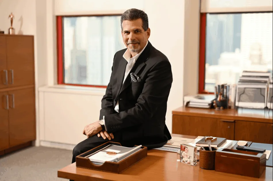 Professional portrait of a businessman in a black suit seated at a desk in a bright office environment.