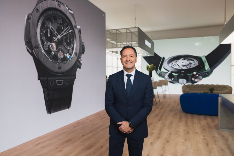 Julien Tornare CEO Hublot 2 Watches World Julien Tornare CEO Hublot Smiling man in a navy suit stands in a watch showroom with oversized watch banners in the background