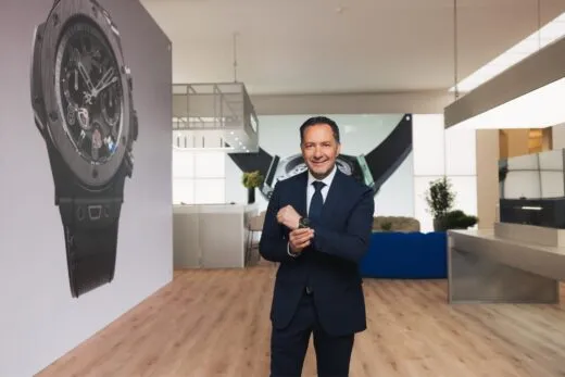 Julien Tornare CEO Hublot Smiling man in a navy suit and tie standing in a bright showroom next to a large wall advertisement of a wristwatch, with wooden floor and modern furniture in the background.