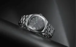 Close-up of a stainless steel wristwatch with a dark dial, date window at 3 o'clock, and a brushed metal bracelet on a black surface.
