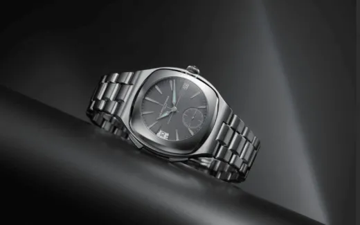 Close-up of a stainless steel wristwatch with a dark dial, date window at 3 o'clock, and a brushed metal bracelet on a black surface.
