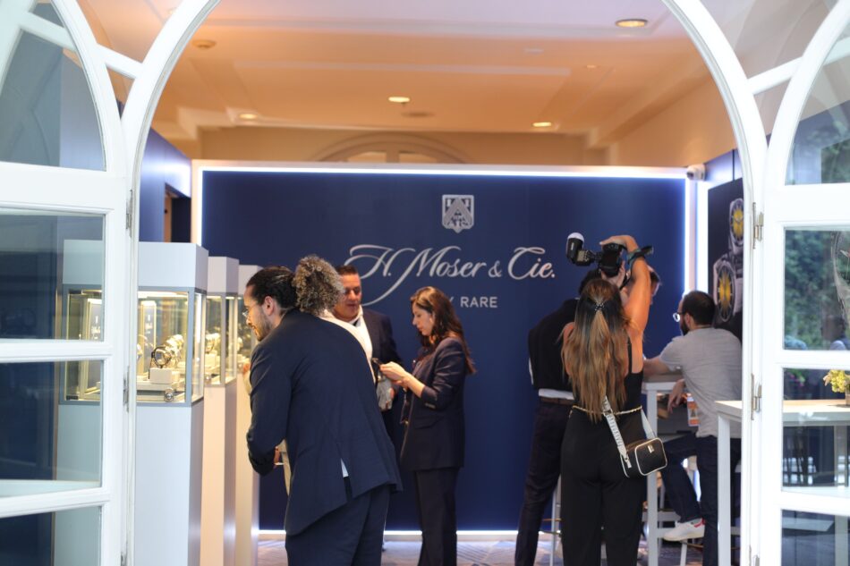 Siar Summer 4 Watches World Crowd of people in formal attire at an exhibition booth with a dark blue branded backdrop and a photographer taking pictures