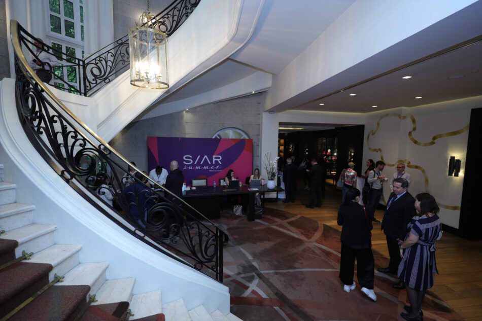 Siar Summer 5 Watches World Elegant hotel lobby with a sweeping curved staircase ornate black railing and a hanging lantern chandelier above a reception area with staff and guests mingling