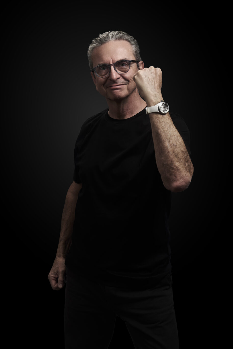 DOMINIQUERENAUD 10 Watches World Portrait of a middle aged man with gray hair and glasses wearing a black shirt fist raised against a dark studio background
