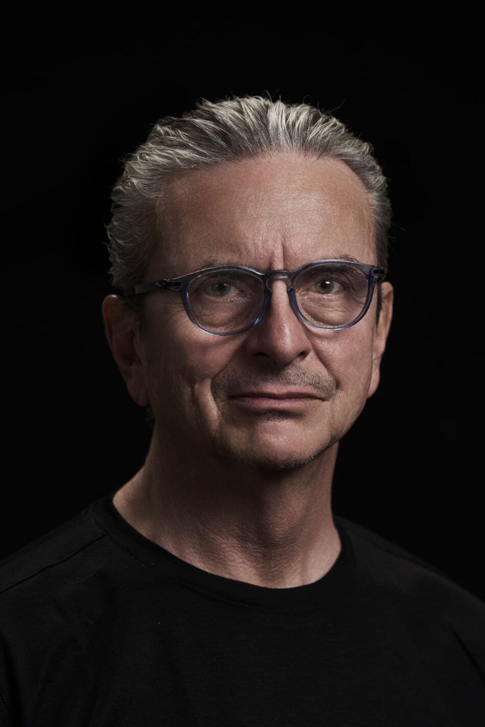 DOMINIQUERENAUD 12 Watches World Close up portrait of a man with gray salt hair and blue rimmed glasses against a black background