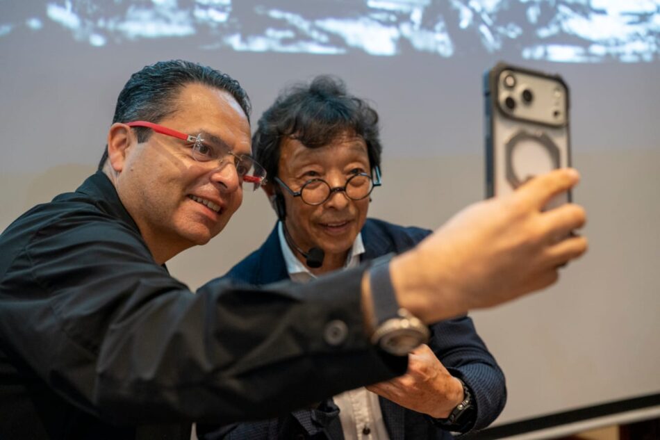 Guillermo Lira y Kiuko Ibe Watches World Two men pose for a selfie with a smartphone smiling at the camera one wears red framed glasses and a headset the other has round glasses and a dark blazer