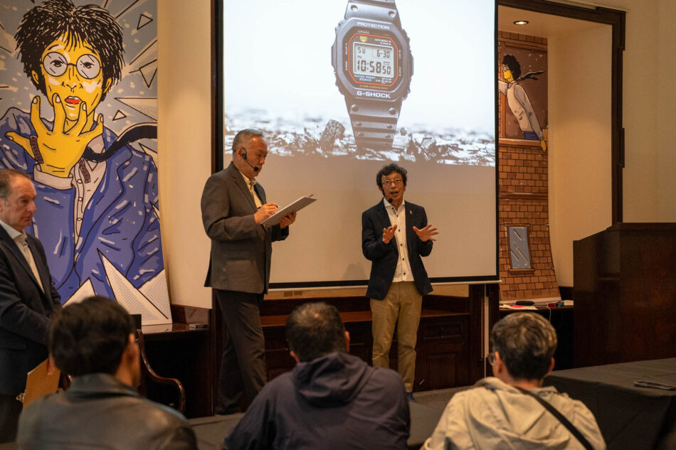 IBE GSHOCK108 Watches World Kiuko Ibe en una sala de conferencias dando una presentación en un escenario otro hombre toma notas en un portapapeles mientras el otro gesticula con la imagen de un reloj gigante proyectada en la pantalla detrás de ellos y el público en primer plano
