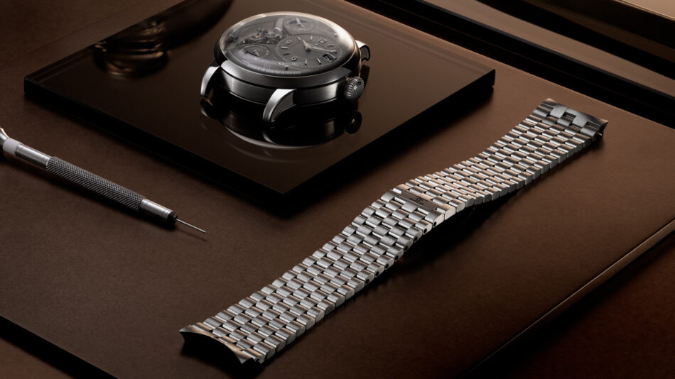 JaegerLeCoultre Duometre Heliotourbillon PerpetualCAJA Y BRAZALETE Watches World Luxurious mechanical watch with exposed gears resting on a glossy black surface beside a precision jewelers tool and a metal watch bracelet on a brown backdrop