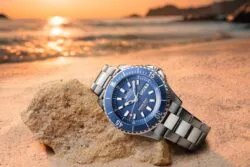 Watch with a blue dial and stainless steel band rests on a sandy rock at sunset beach, showcasing MIDO Ocean Star branding.