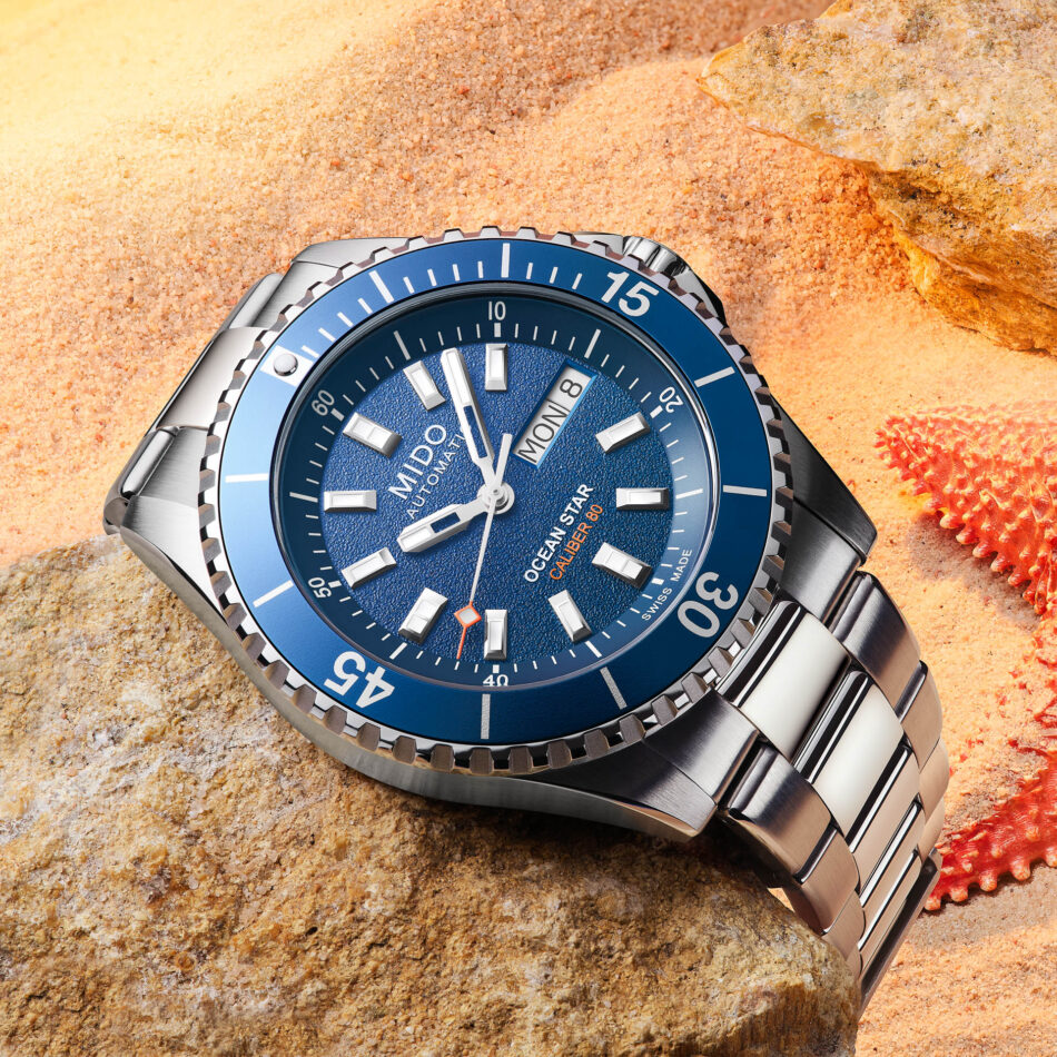 Stacked from 142 images Method=B R=8S=6 Watches World Blue dial stainless steel dive watch resting on orange sand with a rock nearby showing a blue bezel and bracelet pieces