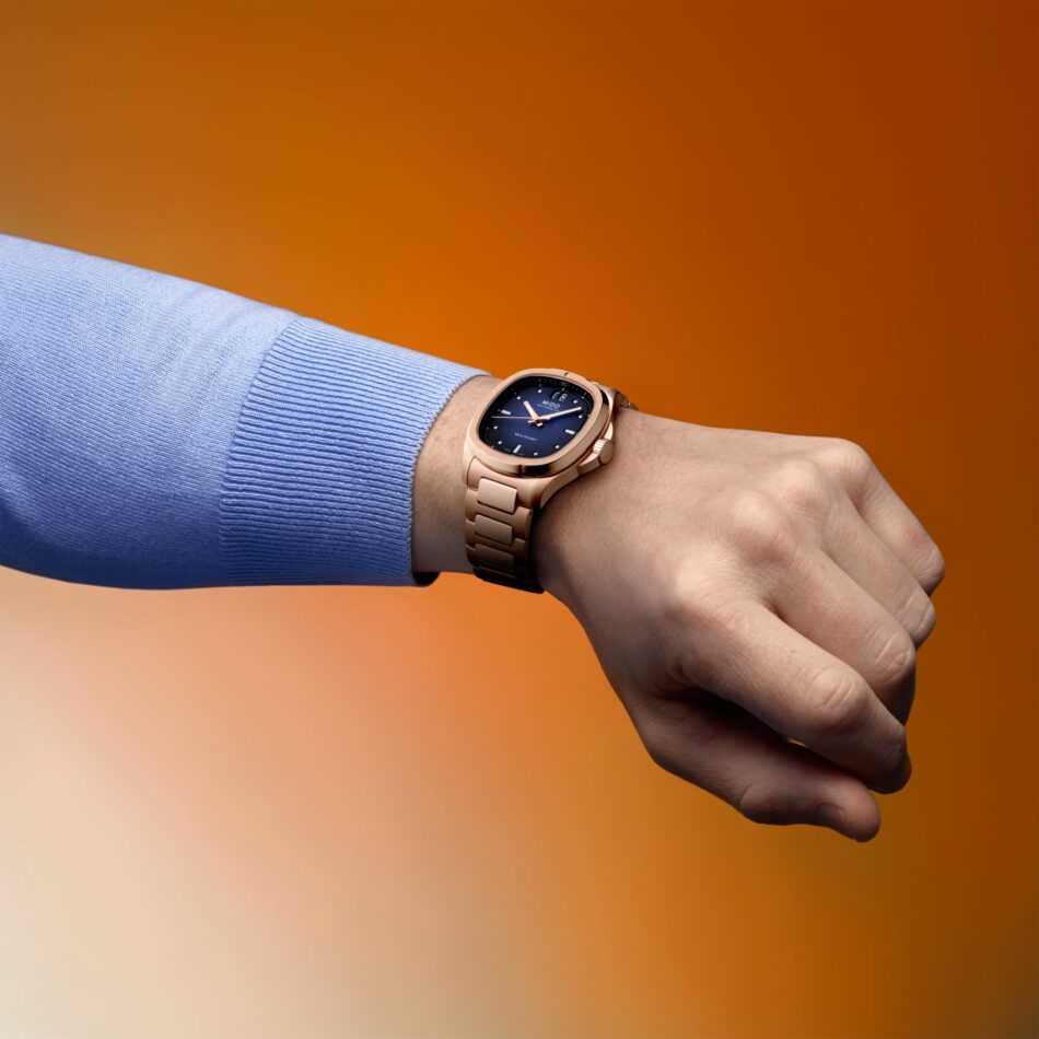 MIDO M0495263304100Multifort TV Big DatePR09 Watches World MIDO Multifort TV Big Date Close up of a rose gold wristwatch on a blue sweater cuff against an orange gradient background