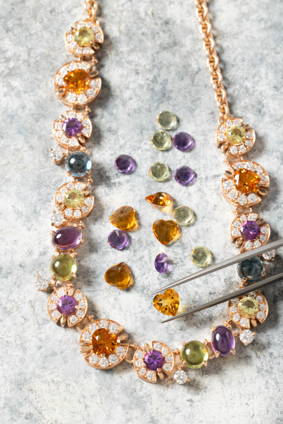 Making of Bvlgari Colors6 Watches World Collar de oro rosa con halos de diamantes y gemas de colores dispuestas sobre una superficie gris rodeado de cuentas sueltas y con unas tijeras cerca