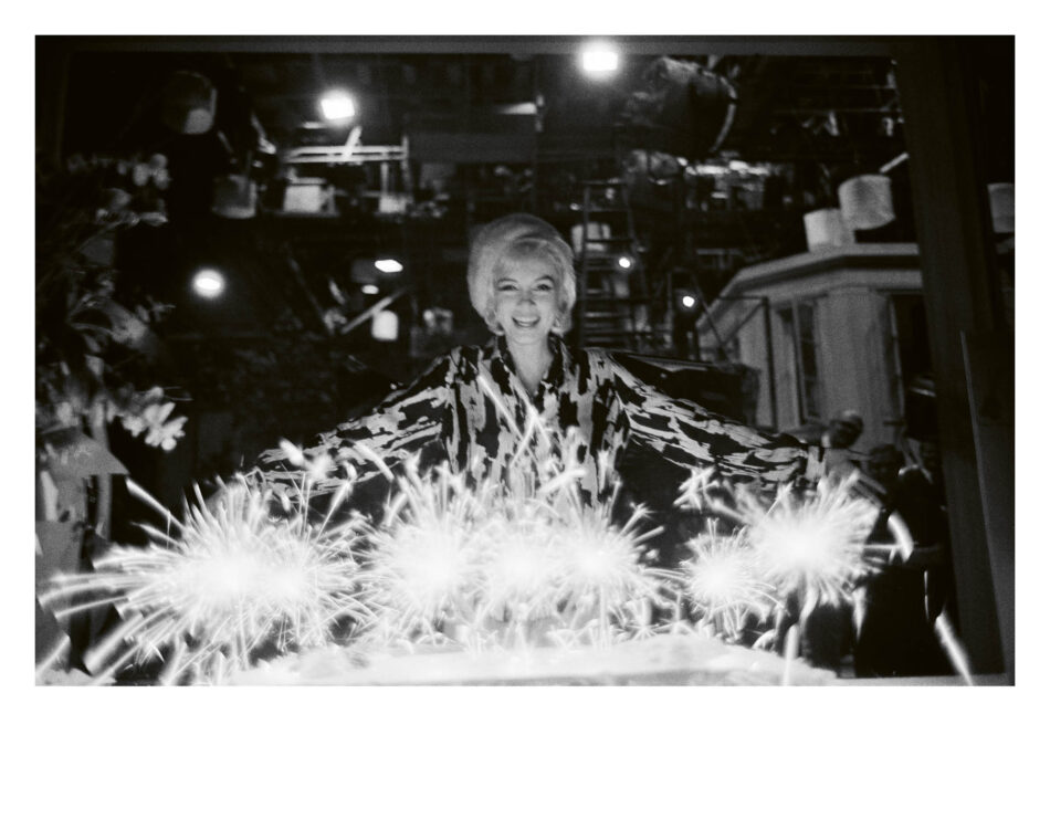 Marilyn Monroe photo by Lawrence Schiller © 2026 Watches World Marilyn Monroe sonriente con una blusa estampada aparece de pie detrás de unos fuegos artificiales brillantes en plena noche en un entorno con poca luz