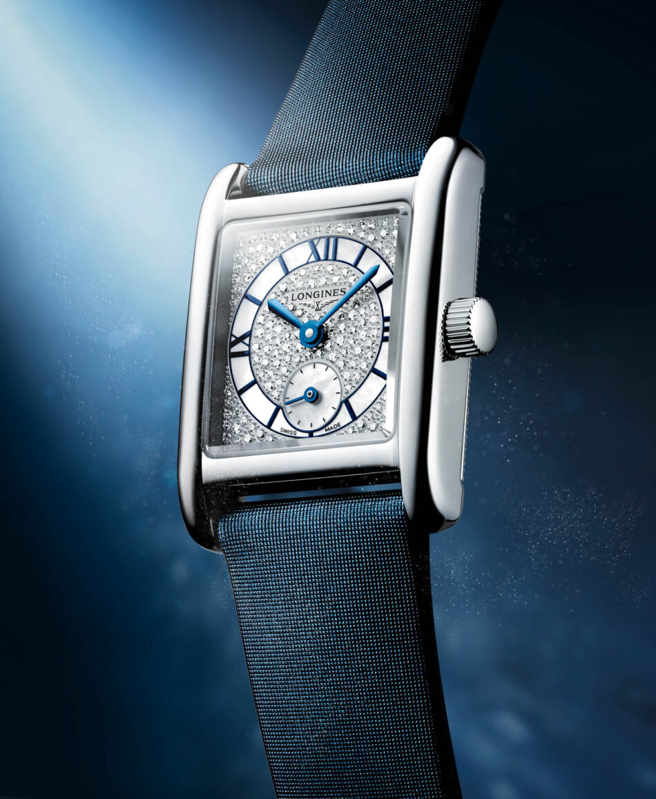 Longines presenta los Mini DolceVita más brillantes y sofisticados hasta la fecha Watches World Close up of a diamond studded rectangular luxury watch with a blue mesh strap against a blue gradient background showing blue hands and Roman numerals on the dial