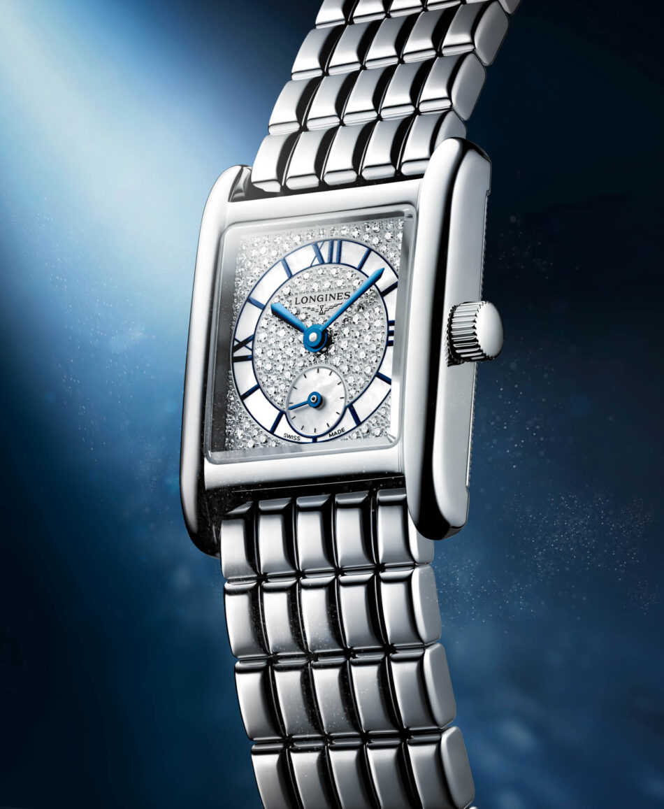 Longines presenta los Mini DolceVita más brillantes y sofisticados hasta la fecha Watches World Close up of a diamond studded Longines wristwatch with a silver bracelet against a blue gradient background angled for depth
