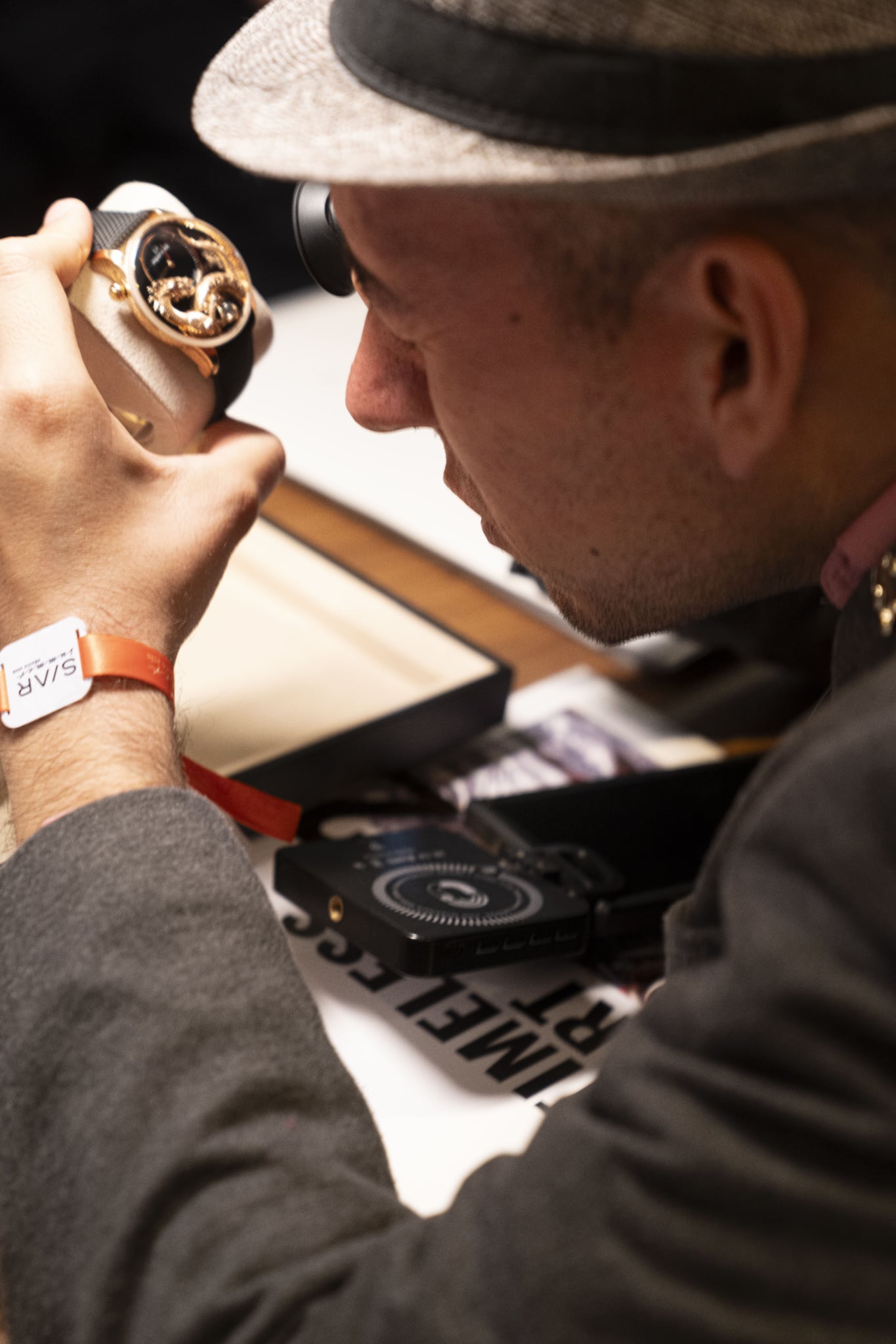 SIAR SUMMER 10 Watches World SIAR SUMMER Close up of a person with a hat inspecting a gold wristwatch using a magnifying loupe seated at a workbench
