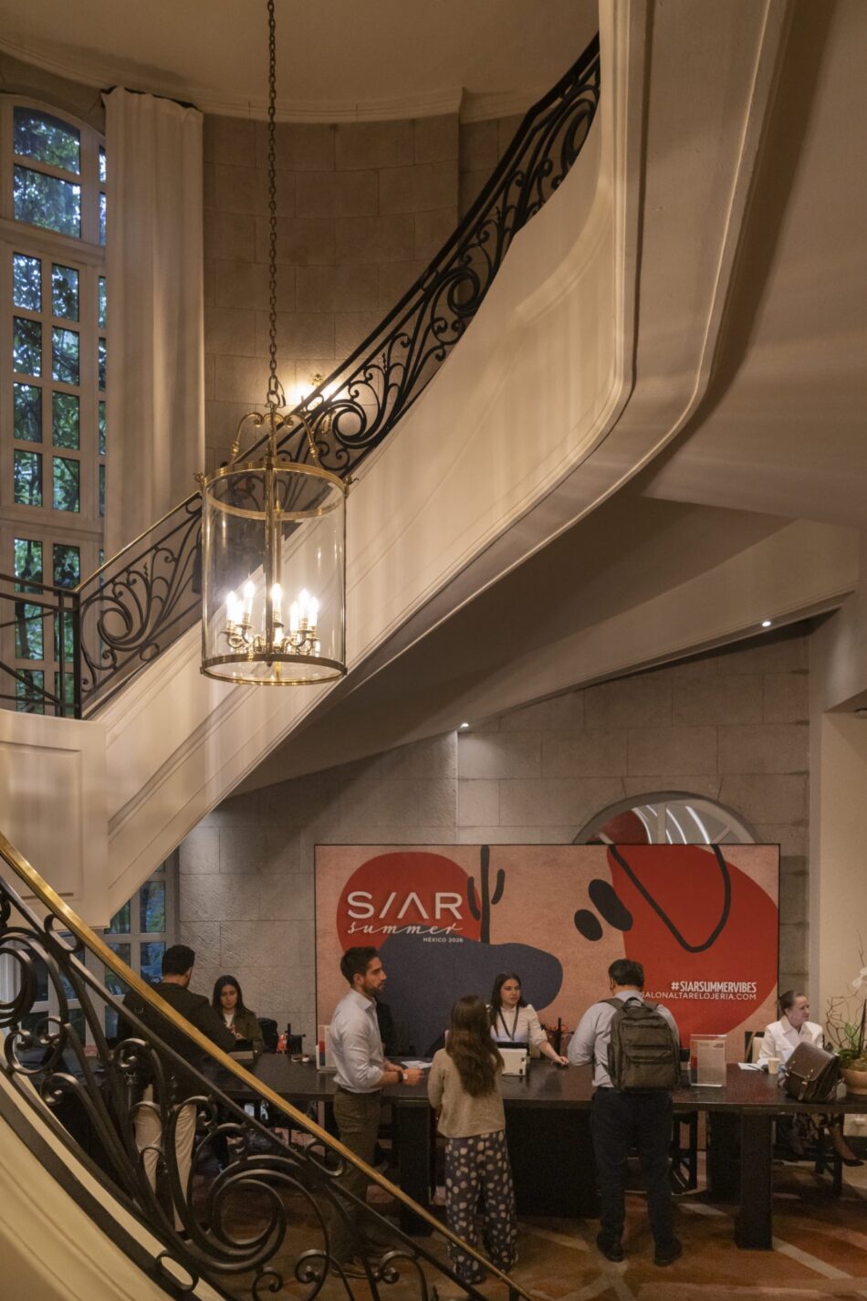 SIAR SUMMER 11 Watches World Curved staircase with ornate black iron railing glass chandelier and people at a reception desk in a grand lobby