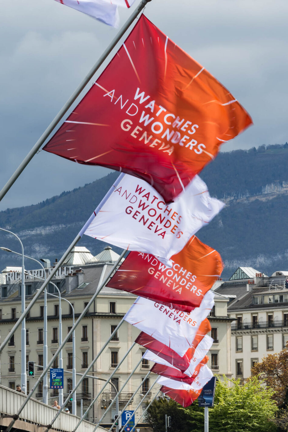SWITZERLAND WATCHES AND WONDERS GENEVA 2026 Watches World Banderas naranjas y blancas con el lema WATCHES AND WONDERS GENEVA ondean a lo largo de una calle de la ciudad con edificios y colinas al fondo