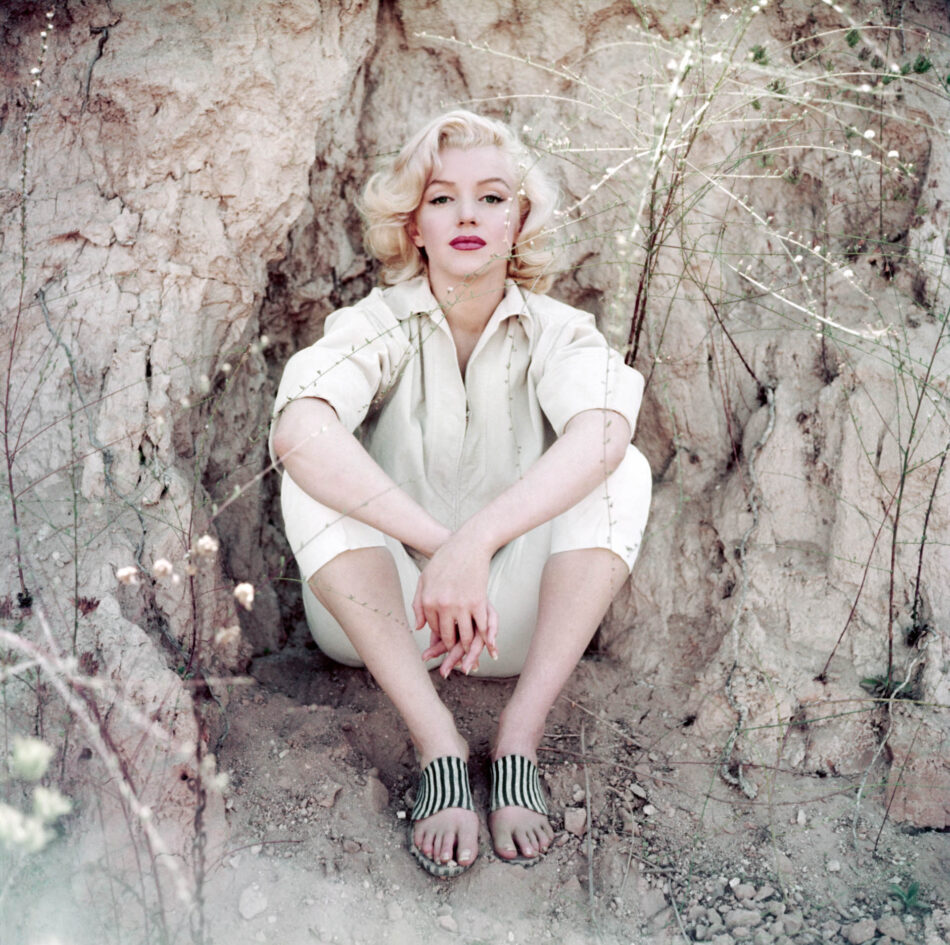XXLPhoto by Milton H Greene © 2026 MHG IP Holdings LLC 8CMYK Watches World Una mujer sentada con las piernas cruzadas entre rocas y hierba seca vestida con un conjunto de color beige claro y calcetines a rayas mirando a la cámara
