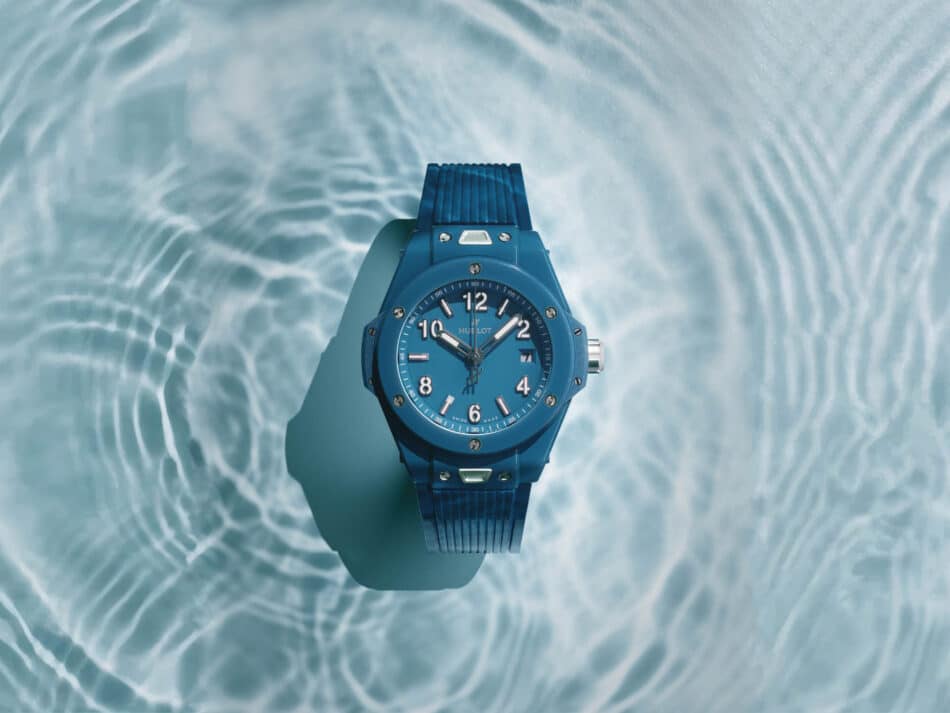 BIG BANG PETROL BLUE CERAMIC 2 Watches World Top down view of a blue wristwatch with a ridged band held over shimmering water ripples