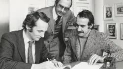 Ferrari TAG Heuer Nikki Lauda Four men in vintage suits inspect sunglasses displayed in a retail case on a store counter. Two men sign documents at a desk while a third man leans over to review, in a vintage office setting.