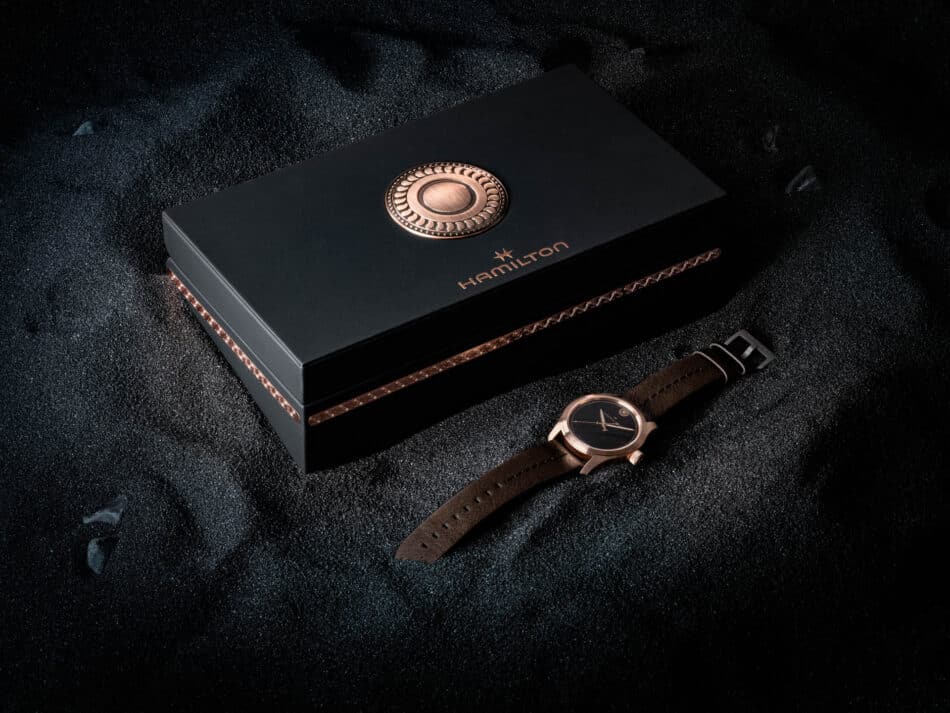 Hamilton Khaki Field Auto The Odyssey Limited EditionH70675530packaging 1 Watches World Black Hamilton presentation box featuring a copper circular emblem on the lid with a brown leather watch strap nearby on a dark textured surface