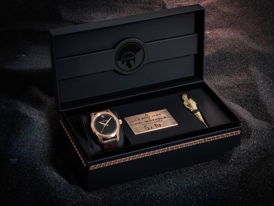 Hamilton Khaki Field Auto The Odyssey Limited EditionH70675530packaging 2 Watches World Black presentation box opened to reveal a gold wristwatch and a bronze plaque set against a dark glittery background