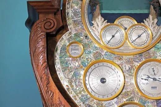 Primer plano de un reloj de madera ornamentado con esferas con bordes dorados e incrustaciones iridiscentes, en cuya esfera se aprecian múltiples indicadores.