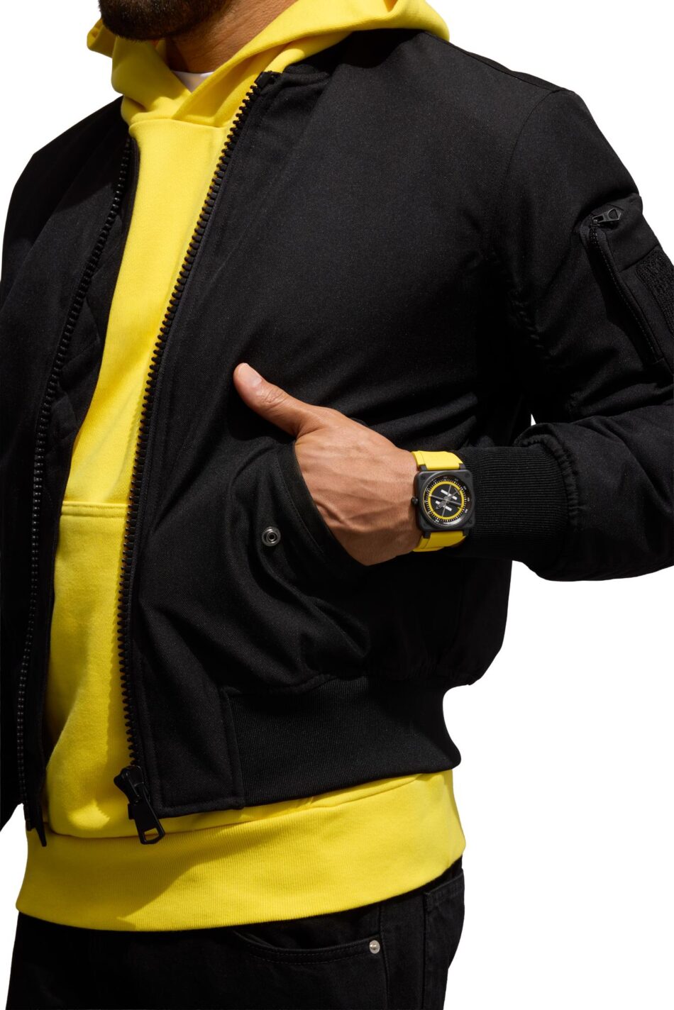 Bell Ross BR03 Helipad Watches World Person wearing a black jacket over a bright yellow hoodie left hand in the jacket pocket yellow wristwatch visible on the wristClose up of a person in a black bomber style jacket with a yellow hoodie underneath yellow watch on the wristWristwatch with a yellow strap on the wearers wrist paired with a black jacket and yellow hoodieBlack jacket with a yellow hoodie and matching yellow watch on the wrist hand tucked in pocketPerson in a neon yellow hoodie under a black zip jacket arm bent with hand in pocket