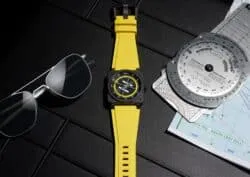 Bright yellow wristwatch with a black watch face on a dark textured surface, beside sunglasses and an aviation chart/map edges nearby.