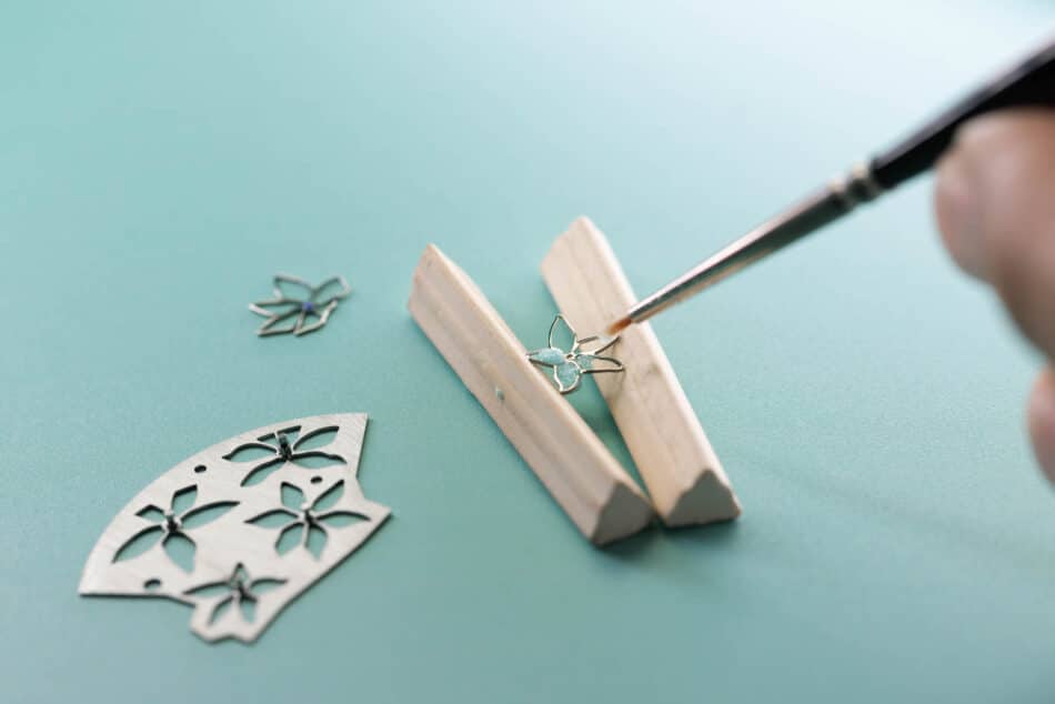 Tiffany Craft Application of the Pliqueàjour Enamel Tiffany Co Watches World Close up of delicate metal flower charms being placed with tweezers on a turquoise craft surface
