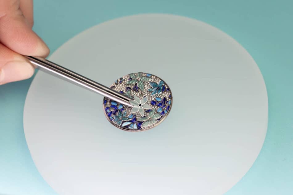 Tiffany Craft Application of the enamel flower on the dial Tiffany Co Watches World Close up of a hand using tweezers to hold a round enamel pendant with blue and green floral motifs against a pale plate and teal background