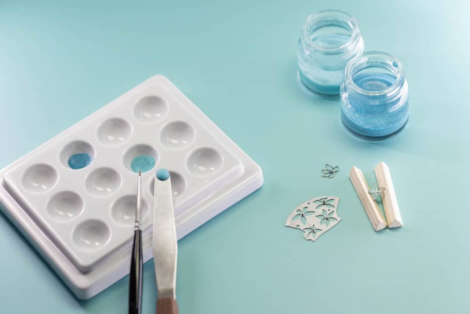 Tiffany Craft The enamel flower 3 Tiffany Co Watches World Jewelry making setup with a white silicone mold blue pigment tweezers and small decorative charms on a pale teal surface